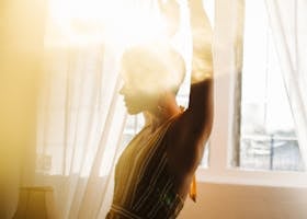 A woman in a striped dress gracefully raises her arms in a sunlit room, exuding peace and inspiration.